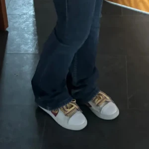 Vita sneakers med färgglada snören och patches - Unika vita sneakers med färgglada, mönstrade snören och broderade patches i olika motiv. Skorna har en klassisk sneakerdesign med rund tå och platt sula. Perfekta för att ge en personlig touch till din outfit.