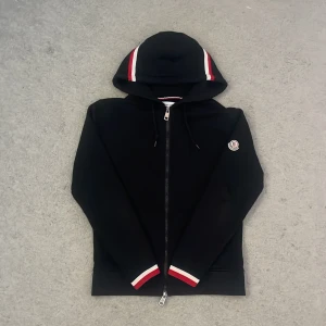 Svart zip hoodie från Moncler - Snygg svart zip hoodie från Moncler med dragkedja och huva. Den har röda och vita detaljer på huvan och ärmsluten. Perfekt för en stilren look. Storlek xxs men är stor i storleken så passar Xs och S. Pris kan diskuteras vid snabb affär.