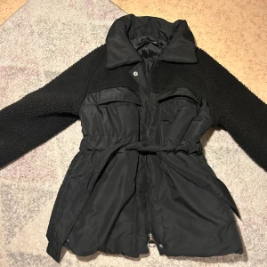 Svart teddyjacka med bälte - Snygg svart teddyjacka med bälte i midjan och dragkedja framtill. Jackan har långa ärmar och en mysig krage. Perfekt för kyligare dagar när du vill hålla stilen.