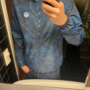 Blå vindjacka med mönster - Säljer en blå vindjacka från  med ett coolt kamouflage mönster. Jackan har dragkedja och huva, perfekt för blåsiga dagar. Den är långärmad och har en normal passform. En stilren och funktionell jacka för alla tillfällen.