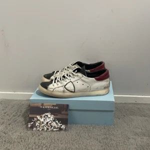 Vita sneakers från Philippe Model - Snygga vita sneakers från Philippe Model med svarta och vinröda detaljer. Skorna har en klassisk snörning och en stilren design som passar perfekt för en avslappnad look.