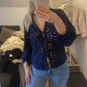 Mörkblå blus med broderade detaljer - Säljer en elegant mörkblå blus med vackra broderade detaljer och knappar framtill. Blusen har långa ärmar och ett genombrutet mönster som ger en unik look. Perfekt för att lyfta vilken outfit som helst!