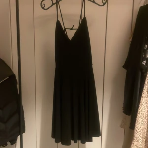 Svart klänning från Bik Bok - Snygg svart klänning från Bik Bok med smala axelband och v-ringad design. Klänningen har en öppen rygg med korsade band, tunt och lätt material som är perfekt att slänga på sig över bikinin på sommaren.