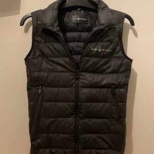 Svart dunväst från Sail Racing - Snygg svart dunväst från Sail Racing med dragkedja och två praktiska fickor framtill. Perfekt för lager-på-lager under kyligare dagar. Diskret logga på bröstet och ryggen ger en sportig touch. Köpt för 1700 stl 150