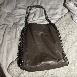 Grå axelväska från Tiamo - Snygg grå axelväska från Tiamo med kedjedetaljer längs kanterna och som axelrem. 