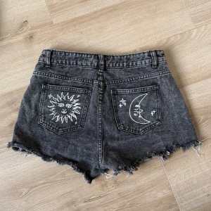 Svarta jeansshorts med sol- och månbroderi - Coola svarta jeansshorts med slitna detaljer och broderier av en sol och en måne på bakfickorna. Shortsen har en fransig kant och en knapp- och dragkedjestängning framtill.