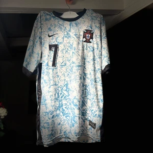 Portugal tröja-Ronaldo 7 24/25 - Säljer en vit blå fotbollströja från Nike med nummer 7 på framsidan och 'Ronaldo' på baksidan. Tröjan har korta ärmar och ett unikt mönster över hela tyget. Perfekt för fotbollsfans som vill sticka ut på planen eller läktaren.