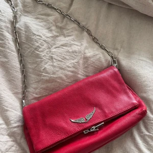 Rocky veske fra zadig - Snygg röd axelväska i skinn med en cool kedja som axelrem. Väskan har en dekorativ silverfärgad detalj framtill och en praktisk dragkedja inuti. Perfekt för att ge din outfit en färgklick!