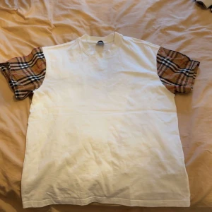 Burberry t shirt - Säljer en stilren vit t-shirt från Burberry med klassiska rutiga ärmar i beige, svart och röd. Perfekt för en trendig look med en touch av lyx. T-shirten har en normal passform och är tillverkad i mjukt bomullsmaterial. Den är L!! Men är liten i storlek så passar M perfekt 