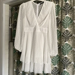 Vit prickig klänning med volangdetaljer - Charmig vit klänning med prickigt mönster och volangdetaljer. Den har en v-ringad design och långa ärmar med elastiska muddar. Perfekt för en somrig look. Gummibandet på ärmen behöver fixas , 