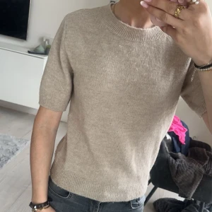 Stickad tröja - Säljer en stilren beige stickad tröja med korta ärmar. Perfekt för en avslappnad look. Tröjan har en rund halsringning och ribbade kanter vid ärmslut och nederkant.