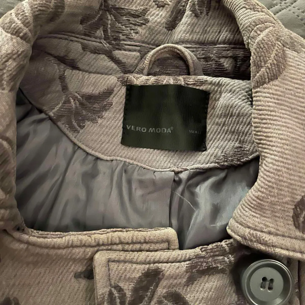 Säljer en elegant kappa från Vero Moda i beige med brunt blommönster. Den har en dubbelknäppt design med stora knappar och långa ärmar. Perfekt för att ge en stilfull touch till din garderob.. Takit.