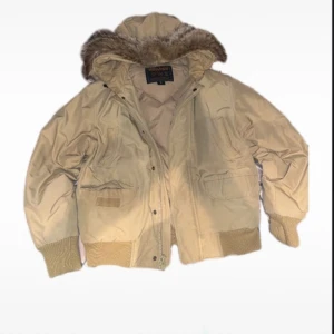 Beige dunjacka från Woolrich - Säljer en beige dunjacka från Woolrich med pälsdetaljer på huvan. Jackan har långa ärmar och ribbade muddar. 