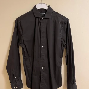 Svart skjorta från Tiger of Sweden - Snygg svart skjorta från Tiger of Sweden i storlek 40. Skjortan har en slim passform och är tillverkad i ett stilrent material. Perfekt för en elegant look med klassiska knappar framtill och långa ärmar.
