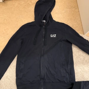 Svart hoodie och byxor från Emporio Armani - Säljer en svart hoodie och matchande byxor från Emporio Armani EA7. Hoodien har dragkedja och långa ärmar med en stilren logga på bröstet. Byxorna har en elastisk midja och logga på benet. Perfekt för en avslappnad stil.