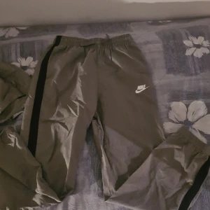 Grå träningsjacka och byxor från Nike - Säljer en grå träningsjacka och matchande byxor från Nike. Jackan har en dragkedja och huva, med svarta detaljer längs ärmarna. Byxorna har en elastisk midja och svarta ränder längs sidorna. Perfekt för sportiga aktiviteter. Oanvända 