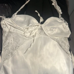 Vit satin klänning med spets detaljer - Elegant vit satinlinne med spetsdetaljer och knytning framtill. Axelbandslös design med vacker spets som ger en romantisk känsla. Perfekt för speciella tillfällen.