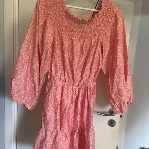 Rosa blommig kortklänning med resår - Söt rosa kortklänning med vitt blommönster och smockad överdel. Klänningen har trekvartsärmar med lätt puff och markerad midja. Perfekt för varma dagar och har en luftig, avslappnad passform.Klänningen är från HM helt oanvänd, med lappen kvar