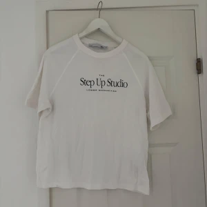 Vit t-shirt Step Up Studio - Vit t-shirt med svart texttryck 'Step Up Studio Lower Manhattan' på bröstet. Klassisk rund halsringning och korta ärmar. Enkel och stilren design, perfekt till jeans eller shorts. Mjuk och skön bomullskvalitet.