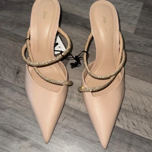 Beige pumps med glittriga remmar från Zara - Snygga beige pumps från Zara med spetsig tå och smala, glittriga remmar i guld över foten. Skorna har en elegant låg klack och är tillverkade i syntetmaterial med en stilren finish. Perfekta för dig som gillar att sticka ut med detaljer. Nypris 599