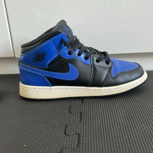 Nike Air Jordan 1 Mid Svart/Blå - Säljer ett par Nike Air Jordan 1 Mid i svart och blått skinn med klassisk vit sula och svarta snören. Ikonisk Jordan-logga på sidan och tungan. Perfekta för dig som vill sticka ut med en tidlös sneaker med streetkänsla. Sulan är lite gul smutsig men syns inte från långt avstånd (ingen låda ) storlek 40/25cm