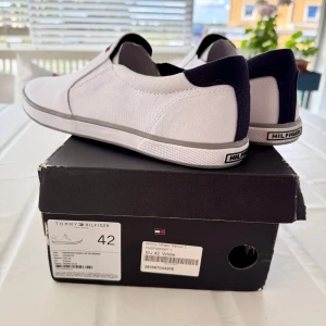 Tommy Hilfiger Vita slip-on sneakers *oanvända* - Snygga vita slip-on sneakers från Tommy Hilfiger med diskret logga på ovansidan. Skorna har en platt sula med grå kant och är tillverkade i textil. Perfekta för en clean och stilren look. Skorna är helt oanvända, kommer med kartong (något sliten kartong) 