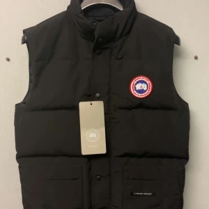 Svart dunväst från Canada Goose - Svart dunväst från Canada Goose med hög krage och klassisk logotyp på bröstet. Västen har två stora fickor framtill och stängs med knappar. Perfekt för lager-på-lager och riktigt snygg till streetwear eller outdoor-stil. 