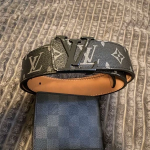 Svart monogram bälte - Snyggt svart bälte från  med klassiskt monogrammönster och stor -logga i metall som spänne. Bältet är gjort i skinn med beige insida och har en exklusiv känsla som lyfter vilken outfit som helst.