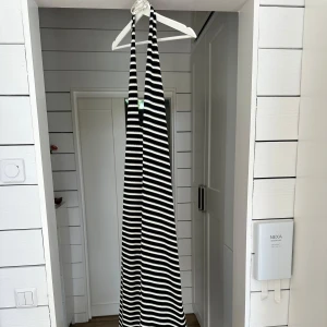 Svartvit randig långklänning - Säljer en svartvit långklänning med smala axelband och djup v-ringning. Klänningen har ett coolt randigt mönster och är tillverkad i ett mjukt, ribbat material som ger en snygg passform. Perfekt för dig som gillar en stilren och trendig look. Tillhörande halssmycke kommer med.