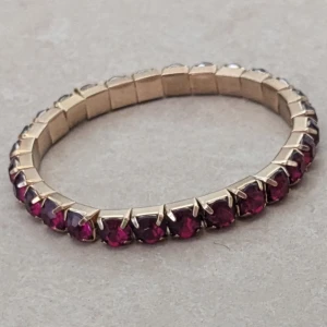 Guldarmband med röda stenar - Snyggt armband i guldton med runda, djupröda stenar runt hela bandet. Klassisk och stilren design som ger en lyxig känsla. Perfekt för dig som vill ha något som sticker ut men ändå känns tidlöst.