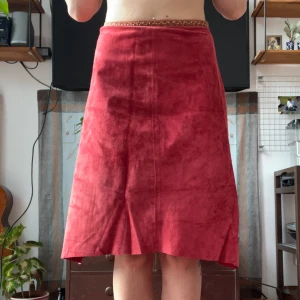 Röd mockakjol från Cam i S - Snygg röd långkjol från Cam i mjuk mocka. Kjolen har en A-linjeformad siluett och ett dekorativt band i midjan med broderad detalj. 