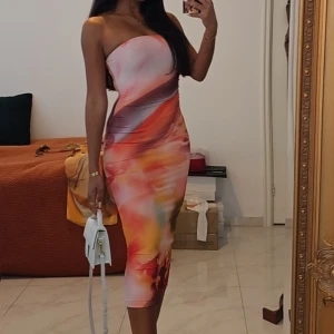 Färgstark axelbandslös långklänning - Säljer en superfin axelbandslös långklänning med färgstarkt abstrakt mönster i rosa, orange, röd och gul. Klänningen har en tight passform som framhäver figuren och är perfekt för dig som vill sticka ut. Materialet känns mjukt och följsamt mot kroppen.