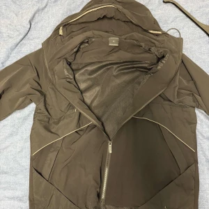 Svart vindjacka från Hoodrich L - Svart vindjacka från Hoodrich med beige detaljer och reflekterande partier på ärmarna. Jackan har huva med dragsko, hel dragkedja framtill och coolt Hoodrich-tryck på bröstet. Perfekt för dig som gillar streetwear och vill ha en lätt jacka med snygg passform.