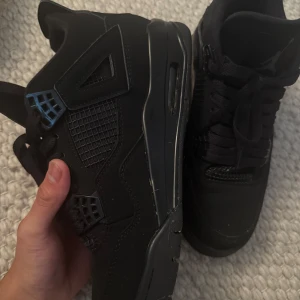 Svarta Nike Air Jordan 4 sneakers - Säljer ett par svarta Nike Air Jordan 4 sneakers med klassisk mesh och läderdetaljer. Skorna har svarta snören, ikonisk Jumpman-logga på plösen och coola plastdetaljer vid snörningen. Perfekta för dig som gillar streetstyle och vill sticka ut.