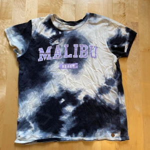 Tie-dye t-shirt med Malibu-tryck - Cool t-shirt från Ü i tie-dye mönster med svart och vit färg. Framsidan har ett stort lila tryck med texten 'MALIBU BEACH'. T-shirten är kortärmad och har en rund hals. Perfekt för dig som gillar avslappnad och trendig stil.