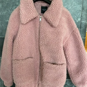 Rosa teddyjacka från New Look Petite - Supermysig rosa teddyjacka från New Look Petite med stora fickor framtill och dragkedja. Jackan har bred krage och är helfodrad, perfekt för kyliga dagar. Den fluffiga looken ger en trendig vibe och passar dig som gillar att sticka ut lite extra.