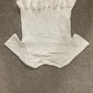 Vit smockad offshoulder topp med volang - Supersöt vit topp med smockad kropp och volang upptill. Offshoulder-modell med korta ärmar och stretchig passform. Perfekt för en fräsch och trendig look till sommaren.