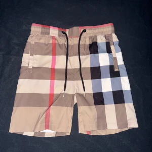 Burberry rutiga shorts med snörning - Snygga shorts från Burberry med klassiskt rutigt mönster i beige, svart, vitt, rött och blått. De har elastisk midja med svart snörning och praktiska fickor fram och bak. Perfekta för sommaren och ger en clean, lyxig vibe till din outfit.