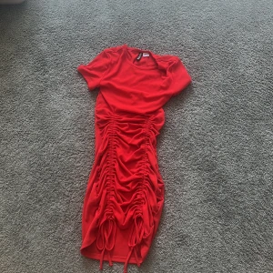 Röd snörad klänning från H&M Divided - Säljer en röd klänning från H&M Divided i storlek XS. Klänningen har korta ärmar, rund hals och cool snörning längs hela framsidan som ger en snygg rynkad effekt. Materialet är mjukt och stretchigt, perfekt för dig som gillar att sticka ut med färg och detaljer.