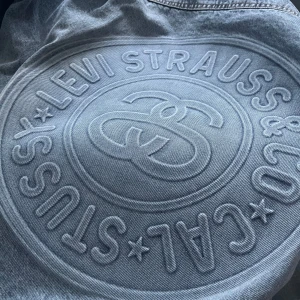 Levi's x Stüssy grå jeansjacka M - Säljer en grå jeansjacka från Levi's x Stüssy med svart krage i manchester. Jackan har stora broderade loggor på ryggen och insidan, samt coolt Stüssy-tryck med 8-ball och ränder på fodret. Klassiska bröstfickor med knappar och relaxed fit.