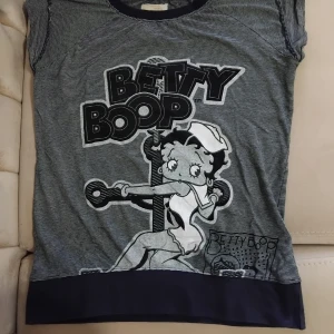 Randig Betty Boop topp Hollywood - Superfin randig T-shirt från Betty Boop x Hollywood med stort tryck framtill av Betty Boop i sjömansstil. Topp med korta ärmar, rund halsringning med knappar på axlarna och mörkblå kant nertill. Perfekt för dig som gillar retro och tecknade motiv.