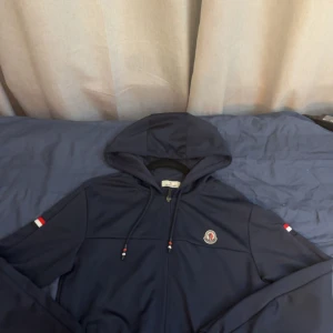 Marinblå vindjacka  Moncler - Snygg marinblå vindjacka Moncler med huva och dragkedja framtill. Jackan har Moncler-logga på bröstet och coola detaljer i rött, vitt och blått på ärmar och snören. Perfekt för dig som gillar stilren och sportig look.