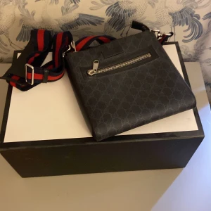 Svart axelväska från Gucci med GG-mönster - Säljer en svart axelväska från Gucci med klassiskt GG-mönster och bred randig axelrem i rött och marinblått. Väskan har en dragkedja framtill och är tillverkad i skinn. Perfekt för dig som vill ha en stilren och lyxig accessoar. Självklart är den äkta och kommer med box, dustbag, kvitto mm, den är köpt i Stockholm för några år sedan för 7500kr! Den har bara legat på en hylla och har knappt blivit använd den är lite dammig eftersom den  inte är använd på länge.