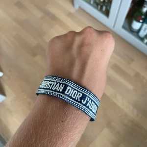 Blått Christian Dior J'Adior armband - Snyggt blått tygarmband från Christian Dior med texten 'J'Adior' broderad i vitt. Armbandet har justerbar knytning med tvinnade band och tofsar i ändarna. Perfekt accessoar för att lyfta din outfit med en lyxig touch.