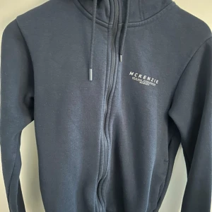 Mörkblå hoodie från McKenzie - Mörkblå hoodie från McKenzie med dragkedja och huva. Tröjan har snörning i huvan och vit logga på bröstet. Tillverkad i mjuk bomullsmix, perfekt för chill dagar eller när du vill ha en enkel streetstyle-look.
