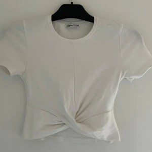 Vit twistad topp från Zara - Säljer en vit kortärmad topp från Zara i storlek S. Toppen har en snygg twistad detalj framtill som ger en cool och modern look. Gjord i mjuk bomull med rund halsringning och figurnära passform. Perfekt att styla med jeans eller kjol.