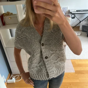 Grå stickad kofta med öppen rygg - Säljer en grå stickad kofta/väst med korta ärmar och snyggt mönster. Koftan har två knappar framtill och har en unik och cool öppning i ryggen!🩷