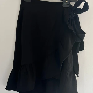 Svart omlottkjol från Vero Moda  - Svart kort omlottkjol från Vero Moda i storlek XS. Kjolen har volangkant nertill och knytdetalj i midjan för en snygg och trendig look. Perfekt för dig som gillar stilrena plagg med en twist.