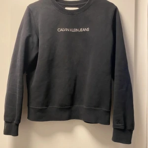 Svart sweatshirt från Calvin Klein Jeans - Svart sweatshirt från Calvin Klein Jeans med vit logotyp broderad på bröstet och diskret CK-detalj på ärmen. Tröjan har rund hals, ribbade muddar och är tillverkad i mjuk bomullsmix. Perfekt för chill dagar och enkel att matcha med jeans eller joggers.