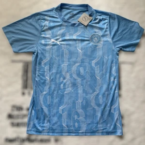 Manchester City blå matchtröja Puma - Snygg Manchester City fotbollströja från Puma i ljusblått med vita grafiska siffror över hela tröjan. Broderat klubbmärke på bröstet och klassisk rund hals. Tillverkad i lätt och ventilerande funktionsmaterial, perfekt för träning eller match.